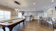 Located at 2605 Elms Plantation Blvd in Charleston, South Carolina, this single-floor community clubhouse is designed to serve as a welcoming social hub for residents. The main gathering area is anchored by a classic wood-framed pool table, positioned beneath a stylish cluster of mason jar pendant lights that add a warm, rustic-modern touch to the space. Hardwood-style flooring runs throughout, contributing to a clean and cohesive aesthetic. A built-in bar counter with bar-height stools provides a casual seating area ideal for socializing, while a wall-mounted flat-screen television enhances the entertainment offerings. Recessed lighting complements the pendant fixtures, ensuring the space is well-lit throughout. Crown molding and a wainscoting-style wall treatment add a refined architectural detail, elevating the overall design. A secondary dining or lounge area with additional high-top seating is visible toward the rear of the space, offering flexible configurations for gatherings. Natural light filters in through a window along the side wall, brightening the interior. The overall ambiance is inviting and functional, thoughtfully designed to encourage community interaction and recreation.