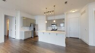 This single-floor residence showcases a thoughtfully designed open-concept layout that seamlessly connects the kitchen and living areas. The kitchen is a standout feature, equipped with sleek gray cabinetry, a white quartz center island with an undermount sink, and a full suite of stainless steel appliances including a French door refrigerator, electric range, built-in microwave, and dishwasher. A marble-look tile backsplash adds a refined touch, while gold-accented pendant and track lighting above the island lends a contemporary warmth to the space. Dark hardwood-style flooring runs consistently throughout the unit, providing a rich, cohesive aesthetic. The bedrooms are bright and well-proportioned, with large windows fitted with white blinds that allow for ample natural light. Each bedroom offers direct access to closet space and connects to one of the two bathrooms. Both bathrooms are finished to a high standard, featuring white quartz countertops, gray cabinetry, large wall mirrors, globe vanity lighting, and tub/shower combinations enclosed by marble-look surround tile with sliding glass doors. The primary bathroom includes a dual-sink vanity, enhancing functionality. Clean white walls and crisp crown molding throughout the unit contribute to a polished, move-in-ready atmosphere.