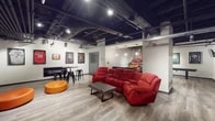 This single-floor sports lounge is designed with a dynamic, industrial aesthetic that sets an energetic and inviting tone throughout. The open-plan layout is anchored by a striking exposed black ceiling featuring visible ductwork, conduit, and track lighting, which lends the space a modern, urban character. Wide-plank wood-look flooring runs throughout, providing warmth and contrast against the industrial overhead elements.

The main lounge area centers around a large red reclining sectional sofa paired with a rustic wood coffee table, creating a comfortable viewing and gathering zone. Two round orange ottomans add a pop of color and flexible seating nearby. A high-top table with black metal stools offers a casual dining or socializing spot. Framed and mounted sports jerseys line the walls, reinforcing the space's athletic theme and adding personality throughout.

A shuffleboard table occupies one area, providing an interactive entertainment option, while wall-mounted flat-screen TVs are positioned for easy viewing from multiple seating areas. A notable architectural feature is the set of full-height glass panel garage doors that connect to an adjacent room or corridor, flooding the space with additional light and offering visual openness. A glass-paneled entry door further enhances the bright, connected feel of the interior.
