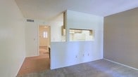This single-floor residence offers a thoughtfully arranged layout that promotes an open, connected feel throughout. The living area is finished with plush carpeting and is anchored by a partial dividing wall featuring a convenient pass-through window that connects directly to the kitchen, allowing for easy interaction between spaces while maintaining a sense of definition. The kitchen is equipped with white appliances, wood-toned cabinetry, and a range hood, providing a functional and tidy cooking environment. A raised countertop along the pass-through serves as an informal bar or serving area, adding versatility to the space. Beyond the kitchen divider, the flooring transitions to a hardwood-style surface, lending warmth and continuity as the layout extends toward additional rooms. A hallway visible from the main living area leads to further private spaces, illuminated by natural light filtering in through a window at its far end. The ceilings feature a textured finish and are designed with slight angles that lend architectural character to the interior. The overall ambiance is clean, bright, and neutral, providing a versatile canvas well-suited for a variety of furnishing styles and personal touches.
