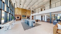 Located at 4126 River Rd in Wilmington, NC, this sophisticated two-story facility offers a generous and thoughtfully designed layout across two well-proportioned floors. The ground floor makes an immediate impression with a dramatic, double-height lobby featuring floor-to-ceiling curtain-wall glazing that floods the interior with natural light and provides sweeping outdoor views. A sculptural undulating ceiling treatment crowns the main gathering area, complemented by statement pendant lighting and large-format porcelain tile flooring. The living room area is anchored by a wood-clad accent wall with a linear fireplace, curated furnishings, and layered lighting that creates a warm yet contemporary ambiance. A dedicated dining area accommodates group gatherings beneath a bold chandelier, while a business support station with cabinetry and print services adds functional convenience. A striking open staircase with steel cable railings and wood treads, framed by a glass curtain wall, connects the floors with architectural elegance. The upper level features a carpeted corridor with an open mezzanine railing overlooking the floors below, leading to four private offices—each finished with designer wallcoverings, built-in desks, and windows with courtyard views. The overall aesthetic is modern and refined, blending clean lines, curated artwork, and warm natural materials throughout.