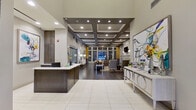 This refined single-floor community space makes a striking first impression with its soaring double-height entry foyer, flooded with natural light through expansive arched windows. A dramatic starburst chandelier anchors the grand entrance, while large-format white tile flooring transitions seamlessly into rich, dark hardwood as you move deeper into the space. The reception area features a sleek, dark-cabinetry desk set against a backdrop of vibrant abstract artwork, lending a polished, contemporary aesthetic. A coffered ceiling with recessed lighting defines the central corridor, adding architectural depth and warmth. The lounge area is generously appointed with plush gray upholstered seating, patterned area rugs in muted gold and charcoal tones, and floor-to-ceiling gridded windows dressed with bold yellow drapery that frame views of an outdoor courtyard. The focal point of the lounge is an elegant white marble fireplace with classical pilaster detailing, topped by a wall-mounted flat-screen television flanked by sconce lighting. Decorative mirrors, curated shelving, and large-scale art pieces throughout add layers of visual interest. The overall ambiance is one of upscale hospitality — bright, inviting, and thoughtfully designed.