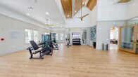 Located on Burbank Avenue in Jacksonville, FL, this single-story fitness center offers a thoroughly equipped exercise facility designed with both functionality and aesthetic appeal in mind. The space is characterized by dramatically high ceilings accented with natural wood beam detailing, white shiplap-style wall panels, and warm-toned hardwood flooring laid in both standard and herringbone patterns throughout.

Large multi-pane windows line the walls, flooding the interior with natural light and providing outdoor views of the surrounding landscape. Ceiling fans are strategically positioned throughout to maintain air circulation across the expansive floor plan. Full-length wall mirrors amplify the sense of space and support proper form during workouts.

The fitness center is divided into distinct functional zones. The strength training area houses multi-station cable machines, adjustable benches, a comprehensive dumbbell rack, and a mounted flat-screen television. A dedicated free weights and functional training zone features kettlebell racks, medicine balls, stability balls, suspension trainers, and a BOSU ball station. The cardio zone is lined with treadmills, stair climbers, and rowing machines positioned alongside the window wall. A tiled accent wall adds a decorative focal point, while built-in cabinetry provides organized storage throughout the space.