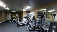 This single-level fitness center offers a practical and well-equipped space designed to support a range of workout needs. The room features a dark ceiling with flush-mounted fluorescent lighting that provides consistent, even illumination throughout. The walls are painted in a warm neutral tone, and a large mirror along one wall visually expands the space while allowing users to monitor their form during exercise. A window fitted with horizontal blinds allows for a degree of natural light.

The cardio section is prominently featured and includes treadmills equipped with individual display screens, an elliptical trainer, and a stationary upright bike. A wall-mounted television is positioned for easy viewing during cardio sessions. On the opposite side of the room, a cable and weight machine offers options for strength training and resistance exercises. The flooring consists of durable, dark carpet suitable for a fitness environment.

Overall, the space is efficiently laid out to maximize equipment availability within its footprint, making it a functional facility for everyday fitness use.