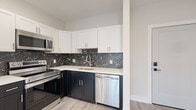This thoughtfully designed single-floor residence showcases contemporary finishes and a clean, modern aesthetic throughout. The kitchen serves as a standout feature, combining crisp white upper cabinetry with deep navy lower cabinets and a striking black marble-patterned tile backsplash. White quartz countertops provide ample workspace, while a full suite of stainless steel appliances — including a French door refrigerator, electric range, built-in microwave, and dishwasher — ensures both style and functionality.

The space includes two distinct bathrooms. One features a full soaking tub set against floor-to-ceiling large-format grey stone tiles, offering a calming retreat. The second bathroom is appointed with a glass-enclosed walk-in shower clad in elegant white marble-look tile with subtle veining, complemented by dark marble-style flooring for a bold contrast.

Living and bedroom areas are finished with light-toned hardwood-style flooring and crisp white walls, creating a bright and airy atmosphere. Recessed lighting adds warmth, while multiple windows with horizontal blinds frame lush, tree-lined views, inviting natural light throughout the day. The overall design balances modern sophistication with comfortable, functional living.