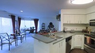 This single-floor residence is designed around an open-concept layout that integrates the kitchen, dining, and living areas into one cohesive, flowing space. The kitchen is outfitted with white cabinetry, a dark countertop peninsula with a built-in stainless steel sink, and a dishwasher. The peninsula serves as both a functional workspace and a visual divider between the kitchen and the adjacent living areas, making it ideal for entertaining or casual dining.

The living and dining areas extend naturally from the kitchen, benefiting from generous natural light provided by large windows dressed with blinds and burgundy curtains. The light-toned carpeting throughout the living space contributes to a warm, comfortable atmosphere. A dining area is positioned near the windows, offering a bright spot for meals.

Decorative elements, including wall clocks, shelving, and greenery placed atop the upper cabinetry, add character to the space. The open sightlines from the kitchen through to the living area create a sense of spaciousness, making the overall layout practical and visually appealing for everyday living.