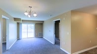 This single-floor space features an open living area finished with wall-to-wall carpet in a cool gray tone, contributing to a clean and understated aesthetic throughout. The room is anchored by a classic multi-arm chandelier with frosted glass shades, providing a focal point overhead while offering ambient lighting. Walls are painted in a warm beige tone, complementing the neutral flooring and creating a cohesive, inviting atmosphere. A large sliding glass door along one wall floods the space with natural light and provides direct access to an outdoor deck, where views of surrounding greenery are visible. A floor-level air vent integrated into the baseboard indicates a central HVAC system. White trim and baseboards offer clean contrast against the neutral wall color throughout the room. A doorway along the interior wall leads to an additional room, suggesting a functional layout with connected living spaces. The overall design is straightforward and well-maintained, with a practical, move-in-ready character suited to a variety of uses. The combination of natural light, outdoor access, and simple finishes gives this space a bright and comfortable feel.