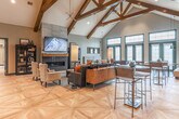 Located at 1709 Cresthill Ave in Royal Oak, Michigan, this single-floor community clubhouse is a thoughtfully designed gathering space that blends rustic architectural character with modern interior finishes. The centerpiece of the space is the dramatic vaulted ceiling adorned with exposed dark wood trusses and beams, which rise steeply overhead to create an expansive, airy atmosphere throughout the main lounge area. Herringbone-patterned hardwood flooring runs throughout, adding warmth and visual continuity across the entire space. The main lounge is furnished with plush sofas and accent chairs arranged around a sleek fireplace topped with a wall-mounted television, offering a comfortable setting for relaxation or socializing. Large floor-to-ceiling windows and transom glass panels along one wall invite abundant natural light while framing outdoor views. A dedicated coffee bar and café-style nook features dark subway tile, open wood shelving, Edison bulb pendant lighting, and a live-edge communal table — creating an inviting spot to gather or work. A game area with a shuffleboard table and intimate seating completes the entertainment offerings. The overall ambiance is modern-rustic, with a carefully curated palette of warm wood tones, muted grays, and deep teal accents that lend the space a refined yet welcoming character.