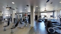 This fitness center offers a thoroughly outfitted workout environment designed to accommodate a wide variety of exercise routines. The space is finished with durable hardwood-style flooring that transitions across distinct workout zones, providing both functionality and a clean, modern aesthetic. Recessed lighting throughout the ceiling ensures consistent illumination, while large windows along the perimeter invite ample natural light and views of the surrounding exterior. Ceiling fans contribute to a comfortable workout atmosphere. The strength training area is well-stocked with a cable machine, adjustable weight benches, free weights, medicine balls, and stability balls, all organized for efficient use. A support column divides the floor plan naturally, creating a sense of separate zones without enclosing the space. On the cardio side, multiple treadmills are positioned near the windows, taking advantage of the natural light. A heavy punching bag is also mounted and ready for use. Mirrored walls on one side enhance the sense of openness and allow users to monitor their form during workouts. The overall ambiance is clean, functional, and well-maintained, making this fitness center a practical and inviting space for a full range of training activities.