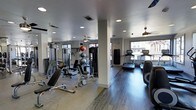 This fitness center offers a thoroughly outfitted workout environment designed to accommodate a wide variety of exercise routines. The space is finished with durable hardwood-style flooring that transitions across distinct workout zones, providing both functionality and a clean, modern aesthetic. Recessed lighting throughout the ceiling ensures consistent illumination, while large windows along the perimeter invite ample natural light and views of the surrounding exterior. Ceiling fans contribute to a comfortable workout atmosphere. The strength training area is well-stocked with a cable machine, adjustable weight benches, free weights, medicine balls, and stability balls, all organized for efficient use. A support column divides the floor plan naturally, creating a sense of separate zones without enclosing the space. On the cardio side, multiple treadmills are positioned near the windows, taking advantage of the natural light. A heavy punching bag is also mounted and ready for use. Mirrored walls on one side enhance the sense of openness and allow users to monitor their form during workouts. The overall ambiance is clean, functional, and well-maintained, making this fitness center a practical and inviting space for a full range of training activities.