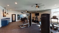 This single-level fitness center is thoughtfully designed to accommodate a comprehensive range of workout styles. The space is divided into distinct functional zones, each outfitted with purpose-specific equipment. The cardio area features True-brand upright bikes, ellipticals, and treadmills, each equipped with individual display screens and positioned near large windows that invite natural light into the room. A wall-mounted flat-screen television is centrally positioned for easy viewing during workouts.

The strength training zone includes a multi-station cable machine, a leg press, and a dedicated free weight area with a tiered dumbbell rack. A large floor-to-ceiling mirror spans one wall, visually expanding the space and providing users with form feedback during exercise. An open functional training alcove, accentuated by a bold navy blue accent wall, houses medicine balls, a weight bench, and additional open floor space ideal for stretching or bodyweight training.

Throughout the facility, durable rubber flooring provides cushioning and traction. Ceiling fans are installed across the space to promote air circulation, and recessed lighting ensures consistent illumination. The overall design is clean and modern, balancing a motivating athletic atmosphere with practical, user-focused layout.