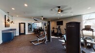 This single-level fitness center is thoughtfully designed to accommodate a comprehensive range of workout styles. The space is divided into distinct functional zones, each outfitted with purpose-specific equipment. The cardio area features True-brand upright bikes, ellipticals, and treadmills, each equipped with individual display screens and positioned near large windows that invite natural light into the room. A wall-mounted flat-screen television is centrally positioned for easy viewing during workouts.

The strength training zone includes a multi-station cable machine, a leg press, and a dedicated free weight area with a tiered dumbbell rack. A large floor-to-ceiling mirror spans one wall, visually expanding the space and providing users with form feedback during exercise. An open functional training alcove, accentuated by a bold navy blue accent wall, houses medicine balls, a weight bench, and additional open floor space ideal for stretching or bodyweight training.

Throughout the facility, durable rubber flooring provides cushioning and traction. Ceiling fans are installed across the space to promote air circulation, and recessed lighting ensures consistent illumination. The overall design is clean and modern, balancing a motivating athletic atmosphere with practical, user-focused layout.