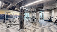 Located at 3601 Market Street in Philadelphia, PA, this fitness center offers a generously sized, single-floor exercise facility designed to accommodate a full range of workout routines. The space features an open, industrial-inspired aesthetic with an exposed ceiling showcasing ductwork and conduit alongside sleek linear LED lighting that keeps the environment well-lit throughout.

The floor is finished with a stylish herringbone-patterned carpet tile in neutral gray tones, adding a modern, polished look to the space. A striking Philadelphia cityscape mural serves as a dramatic focal point along one wall, while floor-to-ceiling glass partitions allow natural light to filter in and provide a sense of openness.

The exercise room is comprehensively equipped with a dedicated cardio zone featuring treadmills, stair climbers, ellipticals, and stationary bikes. A separate strength training area houses a multi-station cable machine, selectorized weight machines, a leg press, and a free weight section with a full dumbbell rack and medicine balls. Large mirrors are strategically placed to maximize the sense of space and assist with form. A built-in storage station near the entrance provides towels and amenities for users. The overall ambiance is clean, modern, and professional, making this fitness center a well-rounded facility for daily training.