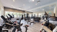 Located at 1601 S Hiatus Rd in Pembroke Pines, Florida, this fitness center offers a thoughtfully designed exercise space that accommodates a comprehensive range of workout needs. The room features durable wood-look flooring, a textured ceiling with recessed lighting, and a ceiling fan that contribute to a clean, modern aesthetic.

Large windows line the walls, flooding the space with abundant natural light while providing views of the surrounding outdoor greenery and pool area. Full-length mirrors along one wall enhance the sense of openness and allow users to monitor their form during exercise.

The cardio section is well-stocked with treadmills, elliptical trainers, stationary bikes, and rowing machines arranged in an organized layout. On the strength side, the space includes a multi-station cable machine, a full dumbbell rack, and various benches to support free weight training. Additional specialized equipment such as recumbent bikes and functional trainers rounds out the facility's offerings.

A wall-mounted flat-screen television adds an entertainment element to cardio workouts. The space is efficiently organized to maximize usability, with clear zones for cardio, strength, and functional training. Ample ventilation and bright, inviting conditions make this fitness center a practical and comfortable environment for residents of the Pembroke Pines community.