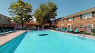 Located at 884 Thurber Dr W in Columbus, Ohio, this outdoor amenity space is centered around a generously sized swimming pool that serves as the focal point of the area. The pool features a bold branded logo on its floor and is filled with clear, vivid blue water, creating an inviting atmosphere for recreation and relaxation. Surrounding the pool is a spacious sun deck outfitted with rows of teal lounge chairs, providing ample seating for sunbathing and outdoor leisure. The deck is enclosed by a sleek metal fence, offering both safety and a defined boundary for the space. The backdrop consists of classic brick residential buildings that lend a timeless architectural character to the setting. Mature, leafy trees frame the perimeter, providing natural shade and a lush, green environment that softens the hardscape. The overall ambiance is that of a well-maintained, community-oriented outdoor retreat designed for warm-weather enjoyment. The combination of vibrant pool water, comfortable furnishings, and verdant surroundings creates a welcoming and visually appealing space that encourages outdoor activity and community gathering.