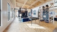 Located at 6601 Roxboro St in Raleigh, NC, this single-story community clubhouse offers a thoughtfully designed, open-concept gathering space with a generous floor area spread across one level. The interior immediately impresses with soaring double-height ceilings in the entry foyer, floor-to-ceiling glass panels, and striking herringbone-patterned hardwood floors that flow throughout the entire space.

The welcoming entryway features decorative floating shelves, a dramatic ring chandelier, and large glass double doors that flood the space with natural light. Adjacent to the entry, a dedicated coffee bar and café-style area is framed by an angular glass and steel clerestory ceiling, patterned tile flooring, a marble-topped island with bar seating, and built-in appliances — ideal for casual gatherings.

The main lounge area showcases exposed wood beam coffered ceilings, a bold dark herringbone tile feature wall anchoring a large mounted television, and comfortable seating arrangements. A foosball table and additional game furnishings make this zone equally suited for recreation. Slatted navy-toned ceiling accents and sculptural globe chandeliers add a modern, upscale ambiance.

A built-in shelving wall divides zones while maintaining visual openness, and a co-working or business center area with workstations is also present. The utility room provides functional support. Abundant windows throughout ensure natural light fills every corner of this vibrant, amenity-rich space.