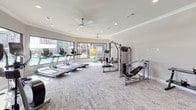 This fitness center in Bryan, Texas is designed to support a comprehensive workout experience within a thoughtfully laid-out, single-floor space. The facility is finished with neutral-toned walls, crown molding, and plush carpet flooring that lends a clean, polished aesthetic throughout. Recessed lighting and multiple ceiling fans ensure the space remains well-lit and comfortable during any workout.

The cardio zone is prominently featured along the windowed perimeter, housing a lineup of treadmills, elliptical trainers, and stationary bikes. Large windows flood the area with natural light while offering open views of the surrounding outdoor amenities, including a pool area and lush landscaping. Full-length mirrors along the walls enhance the sense of spaciousness and provide functional feedback during exercise.

The strength training section is equally well-appointed, featuring a cable machine system, a Smith machine, a multi-function cable crossover unit, and a dedicated free weights area equipped with dumbbells and kettlebells arranged on tiered racks. A flat workout bench and stability balls round out the functional fitness offerings. Bold black interior doors add a striking contrast to the light walls, contributing to the space's modern and inviting character.
