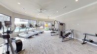 This fitness center in Bryan, Texas is designed to support a comprehensive workout experience within a thoughtfully laid-out, single-floor space. The facility is finished with neutral-toned walls, crown molding, and plush carpet flooring that lends a clean, polished aesthetic throughout. Recessed lighting and multiple ceiling fans ensure the space remains well-lit and comfortable during any workout.

The cardio zone is prominently featured along the windowed perimeter, housing a lineup of treadmills, elliptical trainers, and stationary bikes. Large windows flood the area with natural light while offering open views of the surrounding outdoor amenities, including a pool area and lush landscaping. Full-length mirrors along the walls enhance the sense of spaciousness and provide functional feedback during exercise.

The strength training section is equally well-appointed, featuring a cable machine system, a Smith machine, a multi-function cable crossover unit, and a dedicated free weights area equipped with dumbbells and kettlebells arranged on tiered racks. A flat workout bench and stability balls round out the functional fitness offerings. Bold black interior doors add a striking contrast to the light walls, contributing to the space's modern and inviting character.