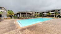 This outdoor pool area is a well-appointed resort-style amenity space designed for relaxation and social gatherings. The centerpiece is a generously sized rectangular swimming pool with crystal-clear blue water, framed by a broad stamped concrete deck that provides ample space for lounging. Rows of chaise lounge chairs line the poolside, offering plenty of seating for residents.

Adjacent to the pool, a built-in outdoor kitchen station features stainless steel grills set into a natural stone surround, making it ideal for outdoor cooking and entertaining. Nearby dining furniture provides a convenient space to enjoy meals al fresco.

A charming wooden gazebo sits at the edge of the pool deck, furnished with wicker seating accented by teal cushions, creating a shaded retreat for relaxing away from the sun. The surrounding landscape is thoughtfully maintained, with manicured shrubs, ornamental grasses, and tall palm trees adding a tropical aesthetic to the environment.

The entire area is enclosed by wrought iron fencing with a gated entry, ensuring privacy and security. A rounded clubhouse building with covered columns and large windows borders one side of the pool, contributing an elegant architectural backdrop. Multi-story residential buildings surround the space, creating a cohesive and well-designed community environment.