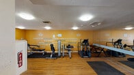 Located at 8111 N 19th Ave in Phoenix, Arizona, this single-floor fitness center is designed to provide a well-rounded workout environment. The space is immediately defined by its bold, golden-yellow accent wall, which is adorned with large-format motivational quotes encouraging a healthy, active lifestyle — including phrases such as "There is no strength where there is no struggle" and "Being healthy isn't a fad — it's a lifestyle."

The room features warm hardwood-style flooring throughout, complemented by a textured white ceiling with flush-mount lighting that keeps the space bright and functional. Full-length wall mirrors along one side visually expand the area and support exercise form checks during workouts.

Equipment includes a selection of strength-training machines, a multi-function cable system, adjustable benches, treadmills, and stability balls, offering options for both cardiovascular and resistance training. Rubber floor mats are positioned beneath the equipment for safety and protection.

Natural light enters through multiple windows fitted with blinds, providing views to the exterior. The space also includes a wall-mounted water fountain and a defibrillator station, reflecting a commitment to member safety and hydration. The overall atmosphere is energetic, clean, and purposefully designed to motivate residents to pursue their fitness goals.