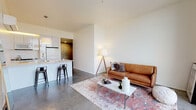 This contemporary single-floor unit showcases a refined industrial-modern aesthetic throughout. The open-plan living area is anchored by striking floor-to-ceiling windows that frame expansive city skyline views, flooding the space with natural light and creating a strong visual connection to the urban landscape beyond. Polished concrete floors run seamlessly throughout, lending a clean, loft-like character to every room.

The living area offers a generous, flexible layout with smooth white walls that serve as a neutral backdrop. It flows naturally into the kitchen, which features a peninsula with bar seating, white cabinetry, light countertops, stainless steel appliances, and a gas range — a functional and well-appointed space suited for everyday cooking.

The bedroom is a calm, minimal retreat accessed via a wide hallway, maintaining the same polished concrete flooring and crisp white walls found throughout the unit. The bathroom presents a compelling contrast of materials, pairing a clean white vanity and quartz countertop with dramatic exposed raw concrete walls, creating a refined industrial character. Vertical backlit mirrors complete the space with a contemporary touch.

Overall, the unit delivers a cohesive, design-forward atmosphere that balances minimalism with warmth, highlighted by abundant natural light and commanding elevated city views.