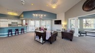 This single-floor community clubhouse offers a thoughtfully designed gathering space with a warm and inviting atmosphere throughout. The centerpiece of the space is a generously sized open lounge area featuring soaring vaulted ceilings accented with bold teal paintwork, creating a striking visual focal point. The lounge is furnished with rich, dark leather sofas and accent chairs arranged around a wall-mounted flat-screen television, set against a decorative patterned wallpapered feature wall. Soft carpet flooring and warm ambient lighting contribute to a relaxed and comfortable environment. Large French doors and multi-pane windows flood the interior with natural light while providing views of the surrounding outdoor area. A set of French doors leads directly into a dedicated fitness room equipped with exercise equipment, offering convenient access for residents. Adjacent to the main lounge is a casual dining nook with a round high-top table and chairs, a coffee station, and informational bulletin boards, creating a functional social hub. A kitchenette bar area with pendant lighting, a tiled countertop, and bar stools provides additional space for informal gatherings. A dedicated workstation area with a desk and chair rounds out the amenities, making this a versatile and well-appointed community facility.