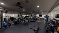 Located at 163 Franklin St in downtown Stamford, Connecticut, this fitness center is a well-appointed, single-floor facility designed to support a comprehensive workout experience. The space features a dark rubber-tile floor that provides both durability and comfort underfoot, while large mirrored walls line the perimeter, visually expanding the room and allowing users to monitor their form throughout their workout.

The cardio zone is equipped with multiple treadmills, elliptical trainers, and a rowing machine, including Precor-branded equipment, catering to a range of aerobic training preferences. Ceiling fans are mounted throughout the space, promoting air circulation during intense sessions, and recessed lighting illuminates the entire floor evenly.

The strength training area offers a well-stocked free weights section with a full rack of dumbbells and kettlebells, a medicine ball tower with various weighted options, and a multi-functional cable/power rack system. Adjustable benches are positioned centrally for versatile use. A wall-mounted flat-screen television is installed in the strength zone, adding an entertainment element to training sessions.

A glass partition separates an adjacent area—visible from the main floor—that appears to house additional amenities. The overall ambiance is modern and focused, with a clean, dark-toned aesthetic that creates an energizing environment suited for both casual and dedicated fitness routines.