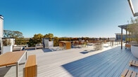 Located at 1880 Maple Dr NW in Kennesaw, Georgia, this expansive rooftop deck offers a thoughtfully designed outdoor gathering space with a variety of distinct zones suited for relaxation, socializing, and al fresco dining. The deck is finished with wide-plank composite decking in a light gray tone, providing a clean and contemporary aesthetic throughout.

Multiple seating areas are distributed across the rooftop, including a covered pergola section supported by bold blue steel posts and fitted with a translucent roof panel, offering shade while maintaining an open-air feel. This sheltered lounge area features plush white upholstered seating and bar-height tables. Elsewhere on the deck, warm-toned teak furniture includes lounge chairs positioned beneath a canopy structure, benches, and bistro-style high-top tables with stools ideal for casual gatherings.

String lights are strung between vertical posts, adding a welcoming ambiance for evening use. Raised planter boxes and large potted greenery add natural softness and visual interest throughout the space. A perimeter railing frames the rooftop, while panoramic views of the tree canopy and distant mountain ridgeline to the north create a striking natural backdrop. The rooftop benefits from abundant natural light and open sky, making it a versatile and inviting outdoor destination.