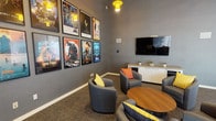 This single-floor cinema-themed lounge is a thoughtfully designed social space that blends entertainment and comfort into a cohesive, visually engaging environment. The room is finished in a deep charcoal-grey palette across the walls, creating a moody, immersive atmosphere accentuated by striking yellow pendant lights that hang from the soaring high ceilings.

A curated gallery of framed classic and contemporary movie posters — including titles such as Star Wars, Guardians of the Galaxy, Godzilla, Wonder Woman, and Creature from the Black Lagoon — lines the walls, serving as the defining decorative feature of the space. A large wall-mounted flat-screen television, flanked by in-wall speakers, is positioned above a light wood and white storage credenza, creating a dedicated media focal point.

The seating area features a cluster of rounded grey upholstered lounge chairs accented with vibrant orange and yellow throw pillows, arranged around a circular wood coffee table to encourage conversation and group viewing. A built-in bar-height counter with black stools along one wall provides additional seating options, complemented by open wall-mounted shelving for decorative accents. Large frosted glass panel partitions along one wall allow soft, diffused natural light to filter into the space while maintaining privacy. Textured grey carpeting grounds the room and adds warmth to the overall design.