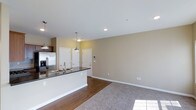 This single-floor residence offers a thoughtfully designed open-concept layout that promotes a natural flow between the kitchen and living areas. The kitchen is a standout feature, anchored by a sleek island with dark countertops and a double basin sink, complemented by stainless steel appliances and dark wood cabinetry. Pendant lighting hangs above the island, adding both style and function to the space. Recessed lighting throughout the ceiling ensures the interior is well-lit and inviting. The flooring transitions smoothly from rich hardwood in the kitchen and entryway to plush carpeting in the living area, creating a warm and comfortable atmosphere. Neutral warm-toned walls provide a versatile backdrop, making the space feel open and airy. Multiple interior doors lead to additional rooms, suggesting a well-organized floor plan with clearly defined private and shared spaces. A wall-mounted smart panel near the entryway adds a modern, connected living touch. Overall, the space combines practical design with contemporary finishes to create a comfortable and functional living environment.