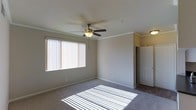 This single-floor residential space is thoughtfully designed with comfort and functionality in mind. The interior showcases wall-to-wall carpeting that adds warmth underfoot, complemented by clean white walls and classic crown molding that frames each room with a refined architectural touch. A ceiling fan with a built-in light fixture is centrally positioned, providing both air circulation and ambient lighting throughout the space. Large windows dressed with vertical blinds allow generous amounts of natural light to flood the interior, casting bright, even illumination across the floors and creating an open, airy atmosphere. The neutral color palette of soft grays and warm whites throughout the space lends a versatile and welcoming feel, making it easy to personalize with various décor styles. Doorways are framed with clean white trim that ties the overall aesthetic together cohesively. The layout flows naturally from room to room, with access to additional areas — including what appears to be a closet or storage space — adding practical utility to the design. Overall, this space offers a clean, well-maintained environment with classic residential features that balance everyday functionality with an inviting and comfortable ambiance.