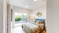 This single-floor space showcases a clean, contemporary design with a cohesive aesthetic carried throughout each room. The bedroom is a serene retreat, filled with natural light that pours in through large sliding glass doors opening to a private outdoor balcony with lush green views. Light hardwood flooring runs throughout, complementing the crisp white walls and adding warmth to the modern palette. A walk-in closet adjacent to the bedroom provides generous storage with open rail systems, keeping the space both functional and organized. A decorative round mirror with an ornate gold frame adds a stylish focal point above the bed, while recessed lighting and bedside lamps create a comfortable, layered ambiance. The suite includes two bathrooms, each thoughtfully designed. The first features a sleek floating vanity with a white quartz countertop, round black-framed mirrors, and warm wall sconces, exuding a boutique hotel-like feel. The second bathroom offers a full soaking tub with floor-to-ceiling neutral tile surround, a white vanity, and warm wood-toned flooring, accented with gold leaf wall décor. Together, these spaces offer a refined, modern living experience with an emphasis on light, comfort, and elevated design details.