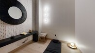 Located at 147 W Wood St in West Lafayette, Indiana, this single-floor wellness suite offers a curated collection of spaces designed for relaxation, mindfulness, and fitness. The layout is compact yet efficiently organized, providing a complete wellness experience within a refined setting.

The entry hallway sets a sophisticated tone with light hardwood flooring, bold terracotta-framed doorways, and a striking accent wall clad in dark panels. A pebbled stone mat at the entryway adds a tactile, spa-like welcome, while a built-in bench with colorful cushions provides a convenient seating area. Cove lighting along the ceiling creates a warm, inviting ambiance.

A dedicated fitness room features floor-to-ceiling windows draped with flowing linen curtains, allowing natural light to filter in while maintaining privacy. Linear LED ceiling lights illuminate the space, which is equipped with a wall-mounted fitness mirror for guided workouts.

The meditation room exudes calm with its textured dotted accent wall, a floating dark console, and three circular sconce lights casting a soft, focused glow. A yoga mat and cushion on the light wood floor make it an ideal space for mindfulness practice.

Completing the suite is a classic sauna finished in natural light wood benches set against dark paneled walls and pebbled stone flooring — a serene retreat for rest and recovery.