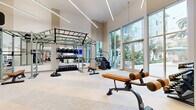 Located at 1001 N Coast Hwy in Oceanside, California, this contemporary fitness center offers a well-appointed, single-floor layout designed to support a comprehensive workout experience. The space is defined by its dramatic soaring ceilings and expansive floor-to-ceiling windows that frame lush palm tree views and flood the interior with abundant natural light, creating an open and energizing atmosphere.

The main workout area is anchored by a large multi-functional rig system equipped with pull-up bars, weight plate storage, medicine balls, and kettlebells, alongside multiple adjustable benches for strength training. A dedicated cardio zone features treadmills, ellipticals, a stair climber, and additional cardio machines, each equipped with individual screens. A wall-mounted display provides an additional media focal point throughout the space.

Supplementary equipment includes a full dumbbell rack, a recovery unit, and plyometric boxes, offering versatility for a wide range of training styles. The light-toned luxury vinyl flooring complements the neutral, warm wall tones and sleek linear LED lighting suspended from the ceiling, contributing to a clean, modern aesthetic. A secondary entrance with double glass doors, a seating bench, and a storage shelf provides a convenient transitional area. The overall design balances functionality with an upscale, resort-like ambiance.