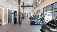 Located in Aiken, South Carolina, this fitness facility offers a thoughtfully designed two-room exercise space that combines modern amenities with an inviting industrial-inspired aesthetic. The main exercise room impresses with soaring ceilings, exposed rustic wood beam columns, and a striking exposed brick accent wall that lends warmth and character to the space. Sleek linear pendant lighting and ceiling fans run along the ceiling, ensuring comfort during workouts. A generous row of large, black-framed windows lines the walls, flooding the floor with natural light and offering views of the surrounding outdoor environment. The rubber flooring throughout provides durability and comfort underfoot. The main room is outfitted with a full lineup of cardio equipment, including treadmills and ellipticals, as well as strength training machines and free weight stations to accommodate a comprehensive workout routine. A secondary exercise room, accessible through a stylish sliding barn-style glass door, provides a more intimate setting ideal for cycling or focused training, and features its own large windows overlooking an outdoor courtyard. Together, the two rooms create a versatile, well-lit fitness environment suited for a wide range of exercise preferences.