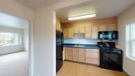 Located at 9061 Seward Park Ave South in Seattle, WA, this single-floor unit offers a practical and well-organized layout suited for comfortable everyday living. The kitchen is equipped with light maple cabinetry, dark countertops, and black appliances including a refrigerator, range, and over-the-range microwave, all set atop warm hardwood-style flooring with a dedicated overhead light fixture for task illumination. The living area is generously sized, finished with neutral carpet and a large window that allows natural light to fill the space, along with a convenient intercom panel near the entry. The bedrooms feature plush carpeting, baseboard heating, and windows that provide views of the surrounding neighborhood. A bright bonus room with hardwood-style flooring and track lighting offers flexible use as a dining area, office, or additional living space. A dedicated utility area houses a stacked washer and dryer alongside closet storage, adding significant convenience. The full bathroom is clean and functional, featuring a soaking tub with shower, a vanity, and grab bars for accessibility. Throughout the unit, a consistent palette of white walls and neutral flooring creates a bright, cohesive atmosphere.