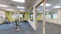 This fitness center is designed to support a comprehensive workout experience across several distinct functional zones. The main exercise area is outfitted with a multi-station cable weight machine, treadmills, a stationary bike, and stair climbers, providing a well-rounded selection of cardiovascular and strength training equipment. Large framed mirrors line portions of the walls, visually expanding the space and allowing users to monitor their form during workouts. The vibrant chartreuse accent walls add energy to the environment, complemented by neutral white walls and cream-trimmed windows and doors throughout.

Natural light floods the space through multiple windows, offering views of lush outdoor greenery and surrounding walkways. The drop-tile ceiling houses recessed panel lighting, ensuring consistent illumination throughout. Colorful, patterned carpet tiles in shades of blue, teal, and gray cover the floors across all areas.

A dedicated open stretching and functional training area provides ample floor space for exercise balls and bodyweight movements. A separate, quieter room features a wall-mounted flat-screen television and windows overlooking the exterior, serving as a media or cool-down space. Direct exterior door access adds convenience for users entering from outdoors.