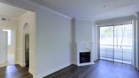 This single-floor space presents a clean, contemporary interior with a neutral color palette and thoughtfully appointed finishes. The main living area is defined by smooth, light gray walls accented by crisp white crown molding, which adds a refined architectural detail along the ceiling perimeter. Underfoot, dark hardwood flooring grounds the space and provides a striking contrast to the bright, airy walls.

A corner fireplace with a white surround serves as a focal point within the living area, offering both visual interest and a functional amenity. Its compact, arched design integrates seamlessly into the room without overwhelming the space.

Natural light is a notable feature, introduced through a large sliding glass door fitted with vertical blinds along one wall. The door provides an outlook to a lush outdoor area, enhancing the sense of openness and connecting the interior to the exterior environment.

Recessed lighting in the ceiling complements the natural light, ensuring the space remains well-lit throughout the day. The overall ambiance is bright, understated, and move-in ready, with a modern yet timeless aesthetic suitable for a variety of furnishing styles.