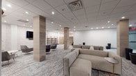 This thoughtfully designed single-floor communal space offers a refined blend of modern aesthetics and functional versatility. The open layout is anchored by distinctive wood-clad structural columns that add warmth and texture against the crisp white walls and light gray patterned carpeting that flows throughout.

The main lounge area features generously sized sectional and modular sofas in neutral tones, arranged to encourage both casual conversation and relaxed lounging. A separate seating zone accommodates accent chairs and an ottoman centered around a wall-mounted flat-screen television, creating a comfortable media area. Recessed lighting in the drop ceiling provides consistent, even illumination across the entire space.

Whitewashed brick-textured accent walls add an industrial-chic character, complemented by open wooden shelving units that offer both display and storage options. A dedicated alcove area includes freestanding wooden privacy dividers with built-in bench seating, ideal for focused, semi-private use.

Toward the rear of the space, a kitchenette with dark cabinetry and a counter-height dining setup serves as a practical refreshment and gathering area. Tall, dark cylindrical planters positioned throughout the corridors add subtle organic accents. Glass-paneled doors connect the space to adjacent areas, maintaining an open and airy feel throughout.