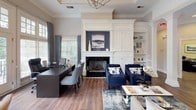 This thoughtfully designed single-floor space exudes classic sophistication through a cohesive blend of architectural detail and refined interior finishes. Rich hardwood flooring runs throughout, anchoring each area with warmth and continuity.

The main living area is a standout, centered around a striking floor-to-ceiling built-in wall unit with integrated cabinetry, open shelving, and a fireplace framed by crisp white millwork. A coffered ceiling with an elegant geometric light fixture adds vertical interest and a sense of grandeur overhead. Navy blue accent chairs provide a bold contrast against the neutral palette, while patterned area rugs layer in texture and depth.

Fluted columns and arched doorways define the transition to a welcoming entryway, where a set of full-glass double doors floods the space with natural light and offers views to the exterior. Crown molding and wainscoting carry throughout, reinforcing the home's architectural character.

A dedicated dining area benefits from floor-to-ceiling windows dressed in bold patterned drapes, allowing ample natural light while maintaining a sense of intimacy. Nearby, a built-in coffee or beverage station with a dark countertop and glass-front cabinetry provides a functional and stylish service nook. A hallway corridor leads to additional rooms, completing this well-appointed and elegantly detailed space.