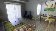 This single-floor unit combines a living area and dining space in an open, functional layout. The living room features durable hardwood-style flooring throughout, complemented by neutral gray walls that create a clean, modern backdrop. A large sliding glass door fitted with vertical blinds floods the space with natural light while providing direct access to an outdoor area, enhancing the sense of openness.

The living area is anchored by a striped multicolor sofa and a dark coffee table, set atop a green area rug that adds a pop of color against the wood-tone floors. A low-profile media console holds a flat-screen television, positioned against the far wall for comfortable viewing.

Adjacent to the living space, a compact dining area is illuminated by a bright window and features a narrow table paired with a yellow metal chair, offering a casual spot for meals or work. The transition between the two areas is seamless, making the space feel cohesive and well-proportioned. Overall, the unit balances practicality with a contemporary aesthetic, making efficient use of the available layout.
