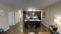This single-floor space showcases a well-designed open-concept layout centered around a modern kitchen that serves as the focal point of the living area. The kitchen features rich, dark espresso-toned cabinetry that extends to the upper walls, providing ample storage while creating a sleek, contemporary aesthetic. Stainless steel appliances, including a refrigerator, electric range, and built-in microwave, are seamlessly integrated into the design.

A peninsula island with a crisp white countertop extends outward, offering both additional workspace and a casual dining area. Two contemporary curved-back bar stools in a warm wood tone are positioned at the island, adding an inviting and functional element to the space.

The flooring throughout is a rich, dark hardwood, which complements the cabinetry and contributes to the cohesive, modern feel of the space. Neutral warm-toned walls provide a versatile backdrop, while track lighting along the ceiling ensures the kitchen and surrounding areas are well-lit. A hallway visible in the background leads to additional rooms, hinting at a thoughtfully connected floor plan throughout the space.