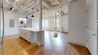 Unit 118 at 5th & Sloan is a thoughtfully designed 2-bedroom, 2-bathroom residence in Columbia, SC, 29205, showcasing an adaptive reuse aesthetic that blends industrial character with contemporary upgrades. The open-concept living and kitchen area immediately impresses with dramatically high exposed ceilings featuring visible steel trusses, ductwork, and structural columns — a hallmark of the building's converted heritage. Warm hardwood flooring runs throughout, complementing the bright, neutral palette of the space. Natural light filters in through large multi-pane windows, enhancing the airy, loft-like atmosphere.

The kitchen is a standout feature, equipped with shaker-style cabinetry, quartz countertops, a subway tile backsplash, stainless steel appliances including a range and built-in microwave, and a generously sized island with pendant lighting above. An in-unit stacked washer and dryer adds everyday convenience.

The spacious primary bedroom features exposed brick walls painted white, hardwood floors, and multiple windows. Both bathrooms are finished with marble-look tile flooring, white shaker vanities, dark countertops, and black hardware accents. One bathroom includes a tub-shower combination with full-height tile surround, while the other offers a window with natural light.