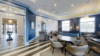 Summerfield Lofts offers a thoughtfully designed community space that balances modern sophistication with welcoming comfort. The single-floor layout centers around a striking lounge area distinguished by a rich navy and white color palette, with deep blue accent walls paired against a textured white brick wall panel that adds visual depth and dimension. A tray ceiling with recessed lighting creates a sense of architectural elegance overhead, while large windows along the perimeter allow natural light to flood the interior, enhancing the airy, open atmosphere. The flooring features a bold, striped pattern in contrasting light and dark tones, grounding the space with a dynamic yet refined aesthetic. Seating arrangements are carefully curated with a mix of plush upholstered chairs, a cushioned sofa, and round tables that encourage both casual conversation and relaxed lounging. A reception-style counter provides a functional focal point near the entrance, making the space versatile for both social gatherings and community activities. Pendant lighting and tasteful wall art add finishing touches that elevate the overall ambiance. The cohesive design of Summerfield Lofts' common area reflects a commitment to providing residents with an inviting, well-appointed environment.