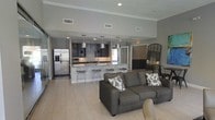 This single-floor space showcases a thoughtfully designed open-concept layout that seamlessly connects the living and kitchen areas into one cohesive, airy environment. The living area is anchored by a plush gray sectional sofa accented with patterned throw pillows, creating a comfortable and stylish focal point. Large-format light tile flooring runs throughout, contributing to the sense of openness and continuity.

The kitchen is a standout feature, boasting dark cabinetry with a sleek finish, dark stone countertops, and a generous kitchen island with bar-height wire seating — ideal for casual dining or entertaining. Stainless steel appliances, including a full-size French door refrigerator, add a polished, professional touch. A trio of modern pendant lights hangs above the island, providing both task lighting and a decorative element.

Crown molding lines the upper perimeter of the space, adding a refined architectural detail, while recessed lighting ensures bright, even illumination throughout. The neutral gray walls provide a versatile backdrop that complements the contemporary design aesthetic. A large arched mirror near the far wall enhances the sense of depth and light. Overall, the space exudes a clean, modern ambiance that is both functional and visually inviting.