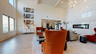 This expansive single-floor space is designed with a refined contemporary aesthetic, combining dramatic architectural elements with thoughtfully curated interiors. Soaring ceilings define the main lounge area, where an abundance of natural light streams through a double-height bank of windows, illuminating light wood-look tile flooring that flows throughout. Bold, large-format abstract artwork in warm amber and slate tones anchors the walls, complementing the rich burnt-orange upholstered seating arrangements that serve as focal points across multiple zones.

A floor-to-ceiling white tile wall features a sleek, linear gas fireplace, creating a striking centerpiece in the main gathering area. Adjacent to this, a generously sized marble-topped island with woven bar stools anchors a bright, open lounge and social space, with floor-to-ceiling windows and glass doors framing outdoor views. A chandelier adds an elegant finishing touch overhead.

A separate dining or co-working area offers built-in banquette booths with mosaic tile backsplashes, dark wood tables, and industrial-style pendant lighting over patterned carpet with orange accent strips. Additional niche seating alcoves with striped upholstered benches and red accent chairs provide intimate gathering spots, completing the space's versatile, upscale ambiance.
