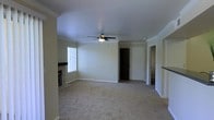This single-floor residence offers a well-organized and comfortable layout with classic design touches throughout. The main living area is generously sized, featuring plush wall-to-wall carpeting, elegant crown molding, and a ceiling fan with a light fixture that adds both function and visual interest. A brick fireplace anchors one end of the room, providing a cozy focal point, while a window allows natural light to fill the space with a warm, inviting glow.

From the living area, an open sightline connects to additional rooms, including a bedroom accessible through a carpeted hallway, maintaining a consistent and cohesive aesthetic. A doorway also leads to a well-appointed kitchen, which is finished with white cabinetry, dark countertops, and stainless steel appliances including a range and over-the-range microwave. The kitchen's tile flooring contrasts neatly with the carpeted living spaces, offering a practical transition between areas.

Throughout the home, neutral wall tones and white trim work together to create a clean, understated atmosphere. Crown molding is carried through multiple rooms, lending a sense of architectural refinement. The overall ambiance is calm and functional, making it a versatile space suitable for a variety of lifestyles.