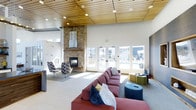 Located at 1420 Renaissance Drive in Longmont, Colorado, this single-floor community space is a thoughtfully designed clubhouse that blends modern aesthetics with warm, inviting elements. The main lounge area immediately draws attention with its soaring ceilings clad in rich wood paneling, which add a natural warmth to the contemporary setting. A striking floor-to-ceiling stacked stone fireplace serves as the room's focal point, anchoring the space with a sense of comfort and rustic character. Recessed lighting throughout ensures the interior remains bright and welcoming. Generous natural light pours in through expansive glass French doors that frame views of the outdoor pool area and surrounding residential community beyond. The lounge is furnished with a bold red sectional sofa, colorful accent seating, and flexible modular ottomans, creating a versatile gathering space suited for both relaxed socializing and casual meetings. Additional seating nooks with accent chairs are positioned near the fireplace, offering a quieter retreat within the larger room. The open layout and abundant glazing create a seamless connection between the interior and the outdoor amenities, making this clubhouse a central hub for community life in this Longmont neighborhood.