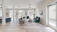 Located at 4201 Runner Loop in Chester, Virginia, this single-story community clubhouse offers a thoughtfully designed gathering space with a bright, contemporary aesthetic throughout. Light wood-look flooring flows seamlessly across the entire layout, complementing the neutral, warm-toned walls and abundant natural light streaming in through oversized windows with transom accents.

The expansive main lounge features soaring ceilings, a ceiling fan, and floor-to-ceiling windows that frame views of the surrounding community, creating an open and airy atmosphere. Comfortable seating arrangements with emerald green and gray upholstered sofas provide inviting spaces for socializing.

A well-appointed kitchen serves as a central hub, showcasing white shaker-style cabinetry with matte black hardware, striking blue herringbone tile backsplashes, white quartz countertops, and a large center island with bar seating. A dedicated coffee bar, also adorned with the signature blue tile, adds a functional and stylish touch.

The dining area features a statement wall with chevron-patterned wood tile, built-in bench seating with blue cushions, and lush tropical plants. A secondary lounge with direct pool access offers pink upholstered seating and views of the outdoor pool deck, blurring the line between indoor comfort and outdoor recreation.