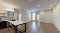 This single-floor unit showcases a contemporary design with a well-thought-out open-concept layout. The kitchen serves as a central hub, equipped with stainless steel appliances including a French door refrigerator, microwave, and range. Cabinetry combines dark espresso lower cabinets with light gray upper cabinets, some featuring frosted glass fronts, all set against a neutral tile backsplash. A freestanding island with a white countertop and dark metal frame provides additional workspace and casual seating. The kitchen flows seamlessly into a generously sized living area finished with warm hardwood-style flooring and recessed lighting throughout.

The bedroom features soft carpeting, a ceiling fan, and a window with exterior views, creating a comfortable retreat. The bathroom is well-appointed with large-format neutral tile surrounding both a soaking tub and a separate walk-in shower. A dark vanity with a light countertop and a wide mirror completes the space. Neutral wall tones and recessed lighting create a cohesive, clean aesthetic throughout the unit, contributing to a bright and modern overall ambiance.